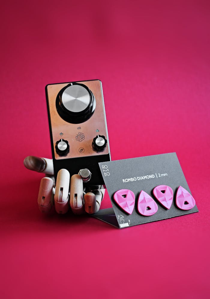 Robotic hand holding guitar picks and audio pedal on vivid pink background.