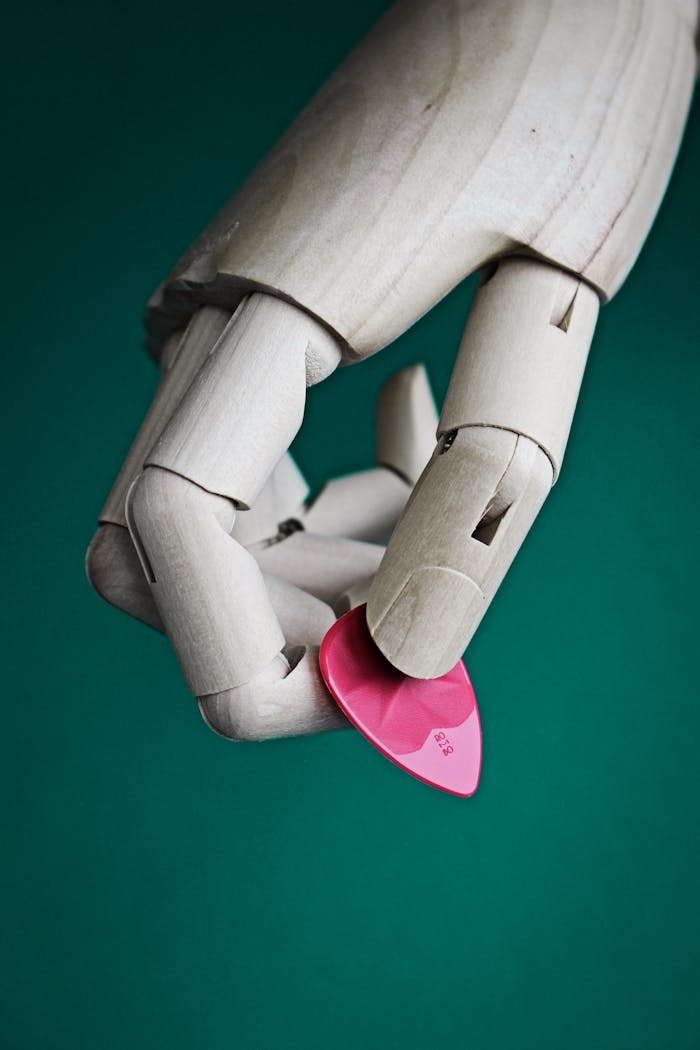 Close-up of a wooden mannequin hand holding a pink guitar pick against a green background.